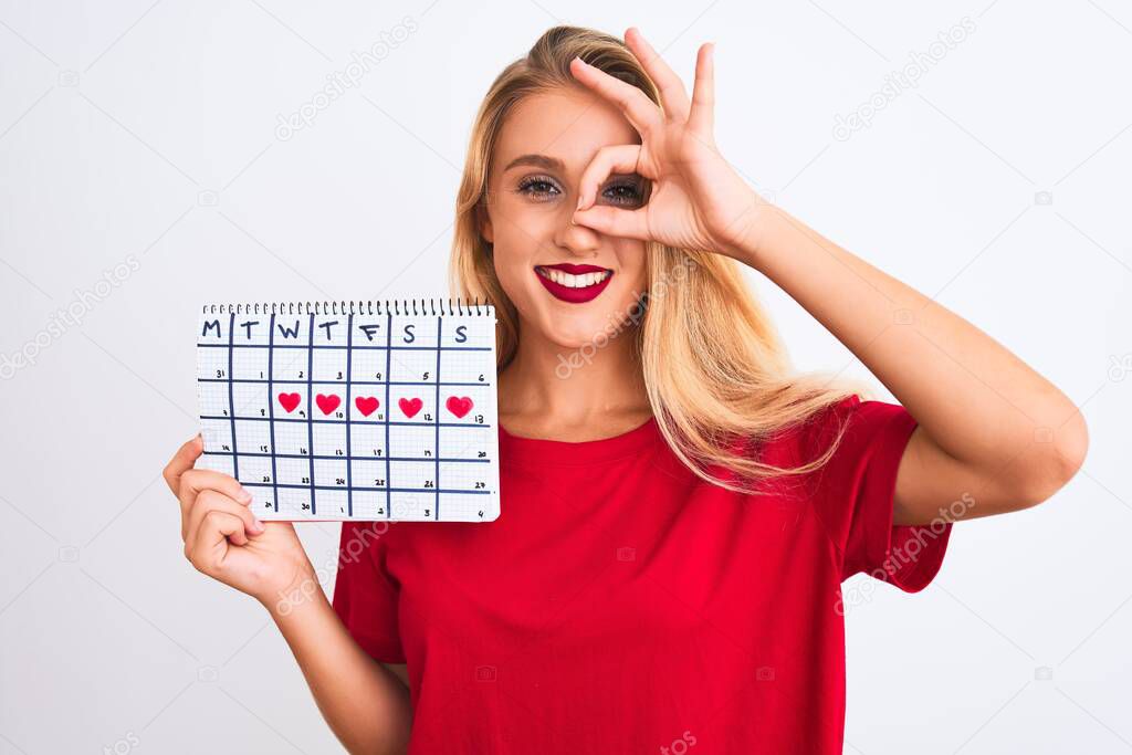 Young beautiful woman holding period calendar standing over isolated white background with happy face smiling doing ok sign with hand on eye looking through fingers