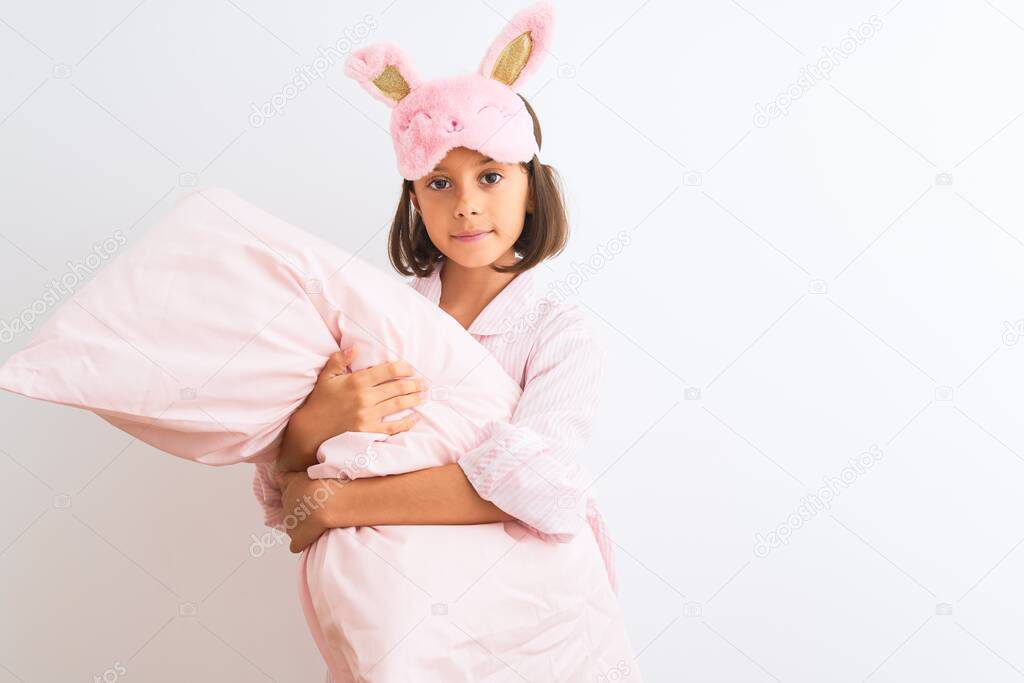 Child girl wearing sleep mask and pajama holding pillow over isolated white background with a confident expression on smart face thinking serious