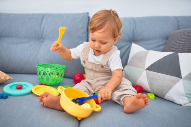 Sevimli sarışın çocuk kanepede oturup evde plastik yemek oyuncaklarıyla oynuyor.