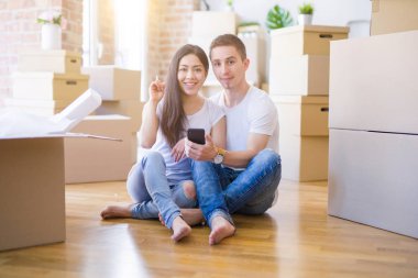 Karton kutuların etrafında akıllı telefon kullanarak yerde oturan güzel bir çift bir fikir ya da soruyla şaşırmış ve mutlu bir yüzle işaret ediyor.