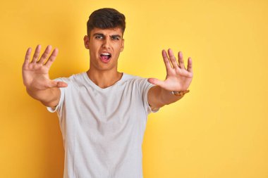 İzole edilmiş sarı arka plan üzerinde duran beyaz tişörtlü genç Hintli adam avuç içi hareketlerini durduruyor, kızgın ve hüsran dolu bir ifade sergiliyor.