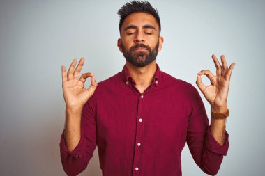 İzole edilmiş gri arka planda duran kırmızı şık gömlekli genç Hintli adam rahatlamış ve gözleri kapalı meditasyon hareketi yaparken parmaklarıyla gülümsüyor. Yoga konsepti.