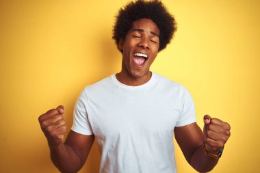 Beyaz tişörtlü, afro saçlı Amerikalı adam izole edilmiş sarı arka planda çok mutlu ve heyecanlı bir şekilde kollarını kaldırıp, gülümseyerek ve başarı için çığlık atarak zafer hareketi yapıyor. Kutlama konsepti.