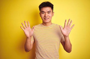 Asyalı genç bir Çinli tişört giyiyor. Sarı arka planda duruyor. Onuncu parmaklarını gösterip gülümsüyor..