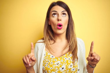Sarı arka plan üzerinde duran genç ve güzel bir kadın şaşırmış ve şaşırmış bir şekilde yukarı bakıyor ve parmaklarıyla işaret ediyor ve kollarını kaldırıyor..
