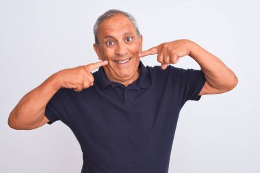 İzole edilmiş beyaz arka planda duran siyah polo giyen yaşlı, gri saçlı, neşeli gülümseyen ve parmaklarıyla ve ağzıyla işaret eden adam. Diş sağlığı konsepti.