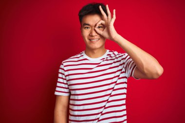 Asyalı genç, çizgili tişört giyen, kırmızı arka planda duran, el hareketleriyle gülümseyen, mutlu yüzüyle parmaklarının arasından bakan Asyalı bir adam..