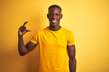 Genç Afro-Amerikalı bir adam günlük tişört giyip izole edilmiş sarı arka planda duruyor gülümsüyor ve el hareketleriyle küçük boy işareti yapıyor ve kameraya bakıyor. Ölçü kavramı.