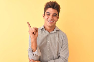 Sarı arka plan üzerinde duran sıradan tişörtlü genç ve yakışıklı bir adam. Yüzünde kocaman bir gülümseme, eli ve parmağı kameraya bakıyor..