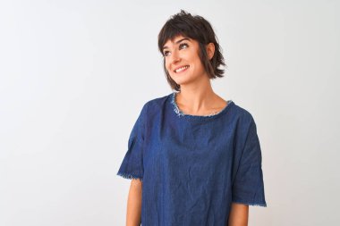 İzole edilmiş beyaz arka plan üzerinde duran kot elbise giyen genç ve güzel bir kadın yüzünde gülümseme, doğal bir ifade ile bir o yana bir bu yana bakıyor. Kendinden emin gülen.