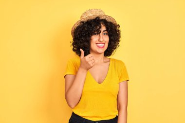 Kıvırcık saçlı, tişörtlü ve şapkalı izole edilmiş sarı arka planı olan genç bir Arap kadın mutlu bir el hareketi yapıyor. Kameraya bakıp başarıyı gösteren ifade onaylanıyor.