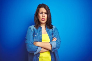Genç, güzel, esmer bir kadın, mavi izole edilmiş arka plan üzerinde sıradan mavi kot ceket giyiyor şüpheci ve gergin, yüzünde çapraz kollu tasvip etmeyen bir ifade var. Negatif kişi.