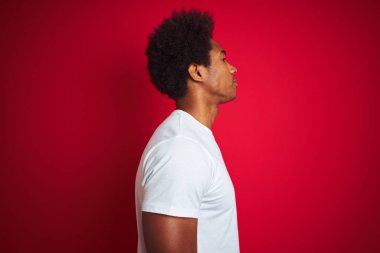 Afro saçlı, beyaz tişörtlü genç bir adam izole edilmiş kırmızı arka plan üzerinde duruyor. Yan tarafa bakıyor, rahat poz veriyor, kendine güvenen bir gülümsemeyle doğal bir poz veriyor..