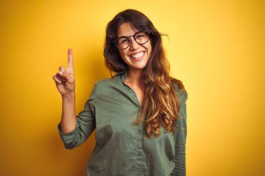 Yeşil gömlek ve gözlük takan genç güzel bir kadın izole edilmiş sarı arka planda bir numaralı parmağı gösterip işaret ederken kendinden emin ve mutlu bir şekilde gülümsüyor..