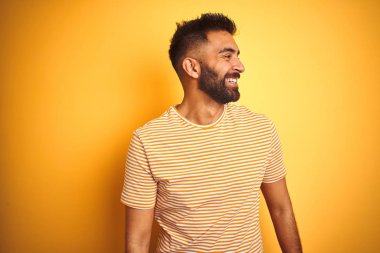 İzole edilmiş sarı arka plan üzerinde duran tişört giyen genç Hintli adam yüzünde gülümseme, doğal bir ifade ile yan yana bakıyor. Kendinden emin gülen.