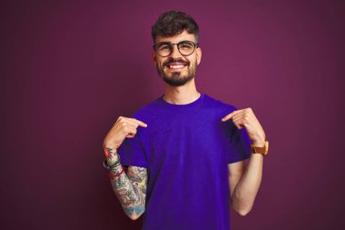Dövmeli, tişörtlü, gözlüklü, mor arka planda duran, kendine güvenen, kendini gururlu ve mutlu gösteren genç bir adam..