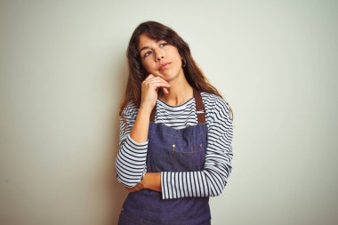 Önlük giymiş genç, güzel bir şef kadın izole edilmiş beyaz arka planda yemek pişiriyor. Elleri çenesinde soru sormayı düşünüyor. Düşünceli bir ifade. Düşünceli bir yüzle gülümsüyor. Şüphe kavramı.