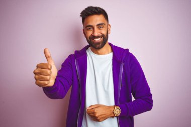 Mor kazak giyen genç Hintli adam pembe arka planda dikiliyor. Gururlu görünüyor, gülümsüyor ve yan tarafa baş parmak işareti yapıyor.