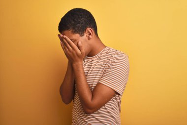 Çizgili tişört giyen genç yakışıklı Arap adam izole edilmiş sarı arka planda dikiliyor ve ağlarken suratını ellerle kapatıyor. Depresyon kavramı.