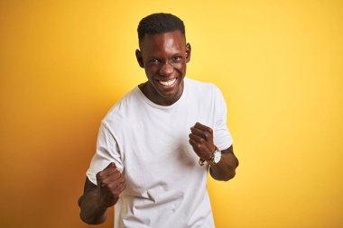 İzole edilmiş sarı arka plan üzerinde duran beyaz tişörtlü genç Afro-Amerikalı adam çok mutlu ve heyecanlı bir şekilde kollarını kaldırıp, gülümseyerek ve başarı için çığlık atarak zafer hareketi yapıyor. Kutlama konsepti.