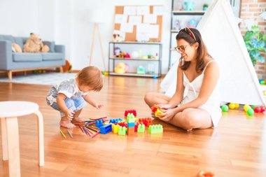 Güzel öğretmen ve bebek anaokulunda bir sürü oyuncağın etrafında yapı taşlarıyla oynuyorlar.