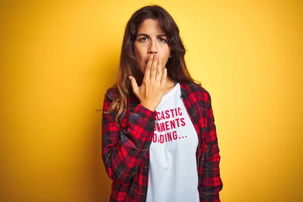 Beautiful woman wearing funny t-shirt with irony comments over isolated yellow background cover mouth with hand shocked with shame for mistake, expression of fear, scared in silence, secret concept