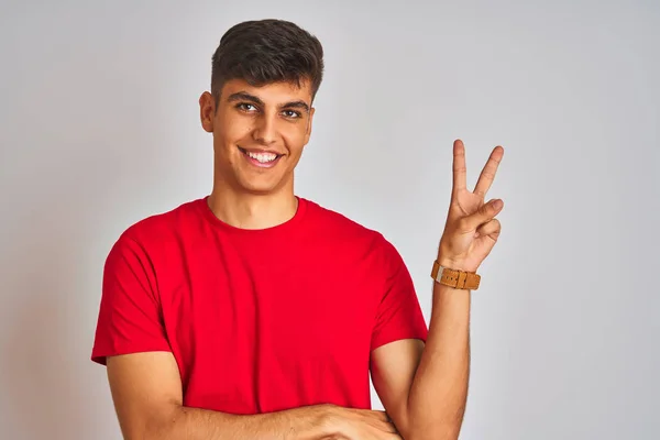 若いインド人男性が赤いTシャツを着て 白い背景の上に立って 幸せな顔をカメラで勝利のサインを行うウィンクで笑っている 第二番 — ストック写真