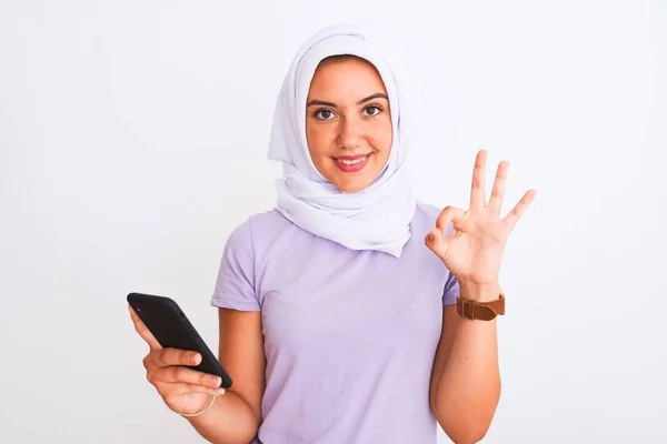 若いです美しいですアラビア語の女の子身に着けていますHijab使用スマートフォン上の隔離された白い背景を持っていますOkサインとともに指 優れたシンボル — ストック写真