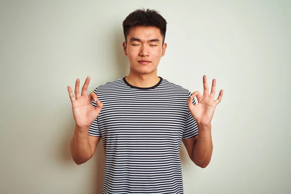 孤立した白い背景の上に立つ縞模様のTシャツを着た若いアジア系中国人男性は 指で瞑想のジェスチャーをして閉じて目でリラックスして笑顔 ヨガのコンセプト — ストック写真
