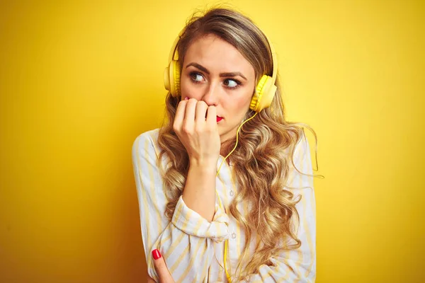 Young beautiful woman listening to music using headphones over yellow isolated background looking stressed and nervous with hands on mouth biting nails. Anxiety problem.