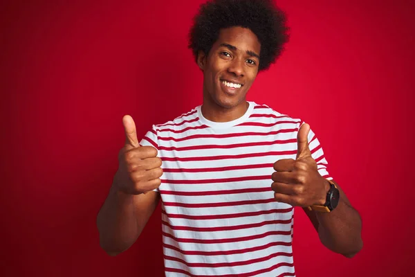 若いアフリカ系アメリカ人の男性は 孤立した赤い背景の成功のサインの上に縞模様のTシャツを着て手で積極的なジェスチャーを行うと 親指を笑顔と幸せ 陽気な表情と勝者のジェスチャー — ストック写真