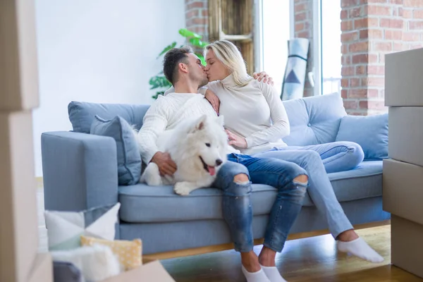 Young beautiful couple with dog kissing sitting on the sofa at new home around cardboard boxes