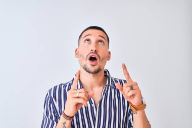 Deniz çizgili gömlek giyen genç yakışıklı arka planda dikiliyor. Şaşırmış ve şaşırmış bir şekilde yukarı bakıyor ve parmaklarıyla işaret ediyor ve kollarını kaldırıyor..