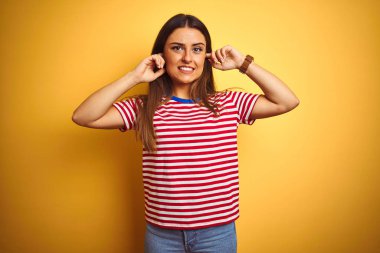 Genç güzel kadın yüksek müzik sesi için rahatsız ifade ile parmakları ile kulakları kapsayan izole sarı arka plan üzerinde duran çizgili t-shirt giyiyor. Sağır kavramı.