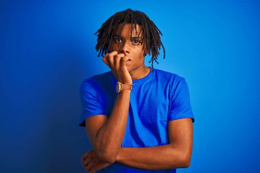 Rastalı tişörtlü Afro-Amerikalı bir adam izole edilmiş mavi arka planda dikiliyor. Elleri ağız ısıran tırnaklarıyla stresli ve gergin görünüyor. Endişe sorunu.