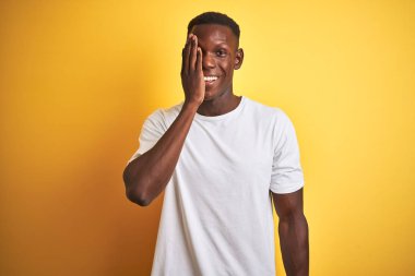 Beyaz t-shirt giyen Afro-Amerikan genç adam izole edilmiş sarı arka planda duruyor. Bir gözünü eliyle kapatıyor. Yüzünde kendine güvenen bir gülümseme ve sürpriz bir duygu var..