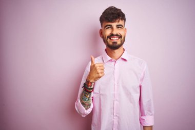 Dövmeli genç bir adam pembe arka planda dikilip el hareketleriyle mutlu bir jest yapıyor. Kameraya bakıp başarıyı gösteren ifade onaylanıyor.