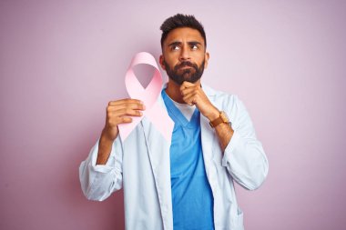 Kanser kurdelesi tutan genç Hintli doktor. Soyutlanmış pembe arka plan üzerinde duruyor. Ciddi bir yüz.