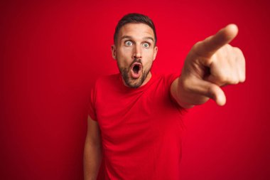 Kırmızı izole edilmiş arka plan üzerine gündelik tişört giyen genç yakışıklı bir adam. Parmakları önde, ağzı açık ve şaşırmış bir şekilde işaret ediyor.