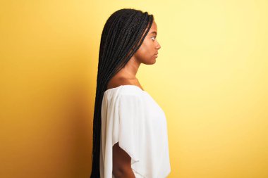 Beyaz tişört giyen Afro-Amerikalı genç bir kadın izole edilmiş sarı arka planda duruyor. Yan tarafa bakıyor, rahat bir görünüm sergiliyor, kendine güvenen bir gülümsemeyle doğal bir yüz ifadesi sergiliyor..