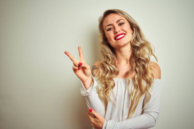 Young beautiful woman standing over white isolated background smiling with happy face winking at the camera doing victory sign. Number two.