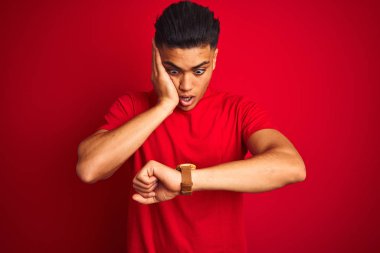 Tişört giyen genç Brezilyalı adam izole edilmiş kırmızı arka planda dikiliyor. Endişeli saate bakıyor, geç kalmaktan korkuyor.