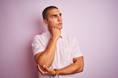 Pembe izole arka planda zarif gömlekler giyen genç yakışıklı bir adam. Elleri çenesinde soru sorarken, düşünceli bir ifade takınıyor. Düşünceli bir yüzle gülümsüyor. Şüphe kavramı.