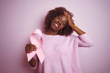 Kanser kurdelesi tutan Afrikalı genç bir kadın pembe arka planda duruyor. Elleri başının üstünde, utanç ve şaşkınlık dolu, sinirli ve hayal kırıklığına uğramış bir halde. Hata için korku ve üzüntü.