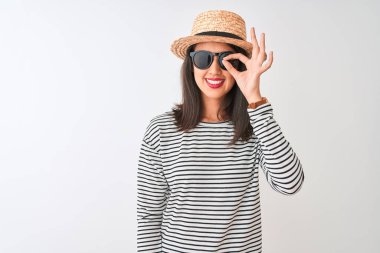 Çinli kadın çizgili t-shirt şapkalı güneş gözlüğü takıyor. İzole edilmiş beyaz arka planda duruyor. El hareketleriyle gülümsüyor..