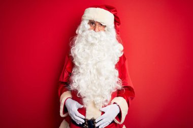 Orta yaşlı, Noel Baba kostümü giyen yakışıklı bir adam izole edilmiş kırmızı arka plan üzerinde duruyor şüpheci ve gergin, sorun yüzünden üzgün. Negatif kişi.
