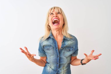Middle age woman wearing casual denim shirt standing over isolated white background crazy and mad shouting and yelling with aggressive expression and arms raised. Frustration concept.