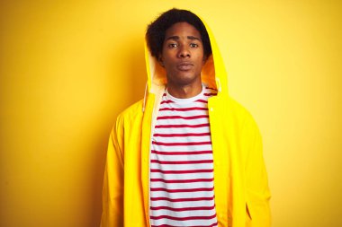 Kapüşonlu yağmurluk giyen afro saçlı Afrikalı adam izole edilmiş sarı arka planda duruyor ve yüzünde ciddi bir ifade var. Basit ve doğal bir şekilde kameraya bakıyorsun..