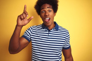 Afro saçlı, lacivert çizgili polo giyen, izole edilmiş sarı arka planda duran Amerikalı bir adam başarılı bir fikri işaret ediyor. Heyecanlı ve mutlu. Bir numara..