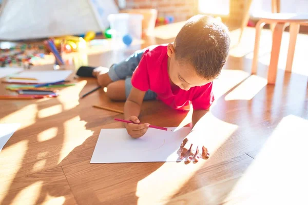 Adorable toddler siting on the floor drawing using paper and pencil around lots of toys at kindergarten